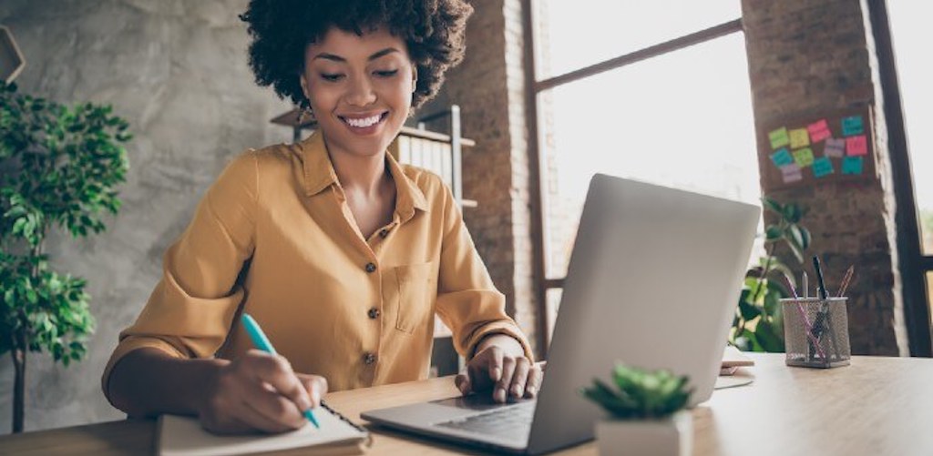 A person smiles and makes a note in front of their laptop